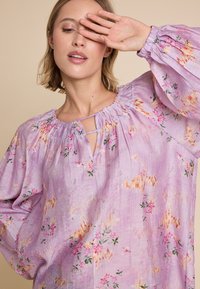 Lilla bluse med blomsterprint i rosa og grønn. Har en rynket halslinning, nøkkelhulldetalj og svevende ermer. Myk tekstur.