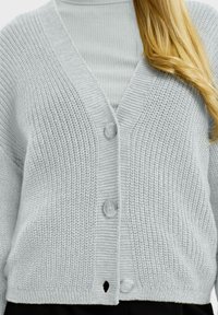 Šviesiai pilkas megztas kardiganas su V formos iškirpte, turintis tris sagas, briaunotas rankoves ir mažą skylutę apvadu.