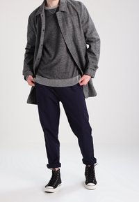 Szary płaszcz w kratę na ciemnoszarym swetrze, granatowych spodniach i czarnych butach za kostkę. Płaszcz ma guziki i kołnierz.