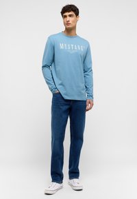 Light blue long-sleeve shirt with “MUSTANG Jeans Company” printed in white. Worn with blue jeans and white sneakers, standing against a gray background.