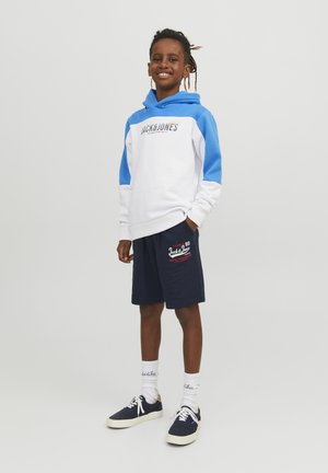 White and blue hooded sweatshirt features front logo, paired with navy shorts displaying red text and graphic. Worn with navy shoes and white socks.