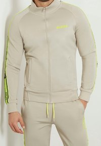 Giacca da track a zip di un beige chiaro con collo alto, strisce laterali giallo neon e dettagli con logo. Tessuto morbido, design aderente e tasche frontali.