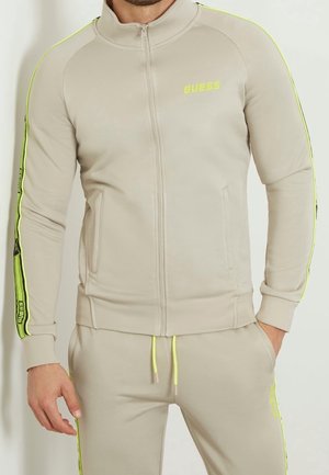 Lichtbeige zip-up sportjack met hoge kraag, neongele zijstrepen en logodetail. Zachte stof, aansluitend ontwerp en voorkruim pockets.