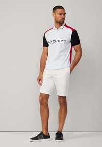 Camisa polo en azul claro con mangas negras y detalles rojos, que presenta un destacado logo de "HACKETT". Combinada con pantalones cortos blancos y zapatillas negras.