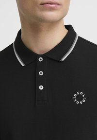 Crna polo majica izrađena od teksturirane tkanine, s kontrastnim sivim ovratnikom, gumbima i okruglim bijelim logotipom na prsima.