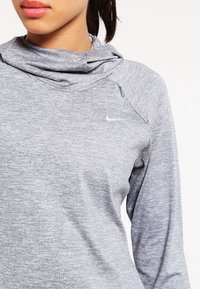 In primo piano, una persona indossa un felpa a maniche lunghe con cappuccio grigia della Nike, con una piccola cerniera sulla spalla e il logo swoosh ben visibile.