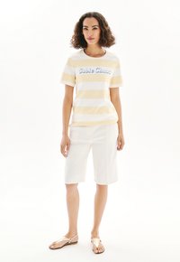 Randig T-shirt med kort ärm i ljungul och vit, med blå texten "Sable Chant" över bröstet, matchad med vita knälånga shorts och guld sandaler.
