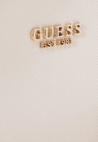Biała teksturowana powierzchnia z złotym logo "GUESS" oraz detalem "EST 1981", prezentująca wytłoczone wzory i gładkie wykończenie.
