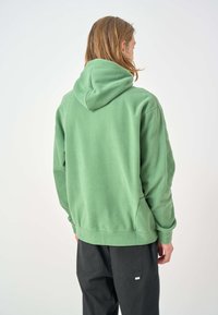 Green hoodie made of soft material, featuring a hood, kangaroo pocket, and ribbed cuffs. Model wearing dark pants, shown from the back.