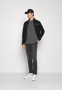Sort ruskindsjakke med lynlåsdetaljer, grå polo-shirt, mørke jeans og hvide sneakers. Modellen bærer en sort kasket.