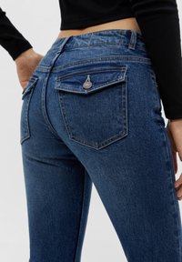 Jeans de mezclilla azul oscuro con corte ajustado. Presentan dos bolsillos traseros con detalles de botón y una textura suave. Costuras visibles.