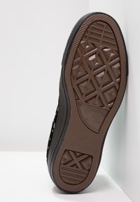 Svart sneaker med en strukturerad gummisula som har ett diamantmönster och varumärkessymboler. Ovandelen har cirkulära utskärningar för ventilation.