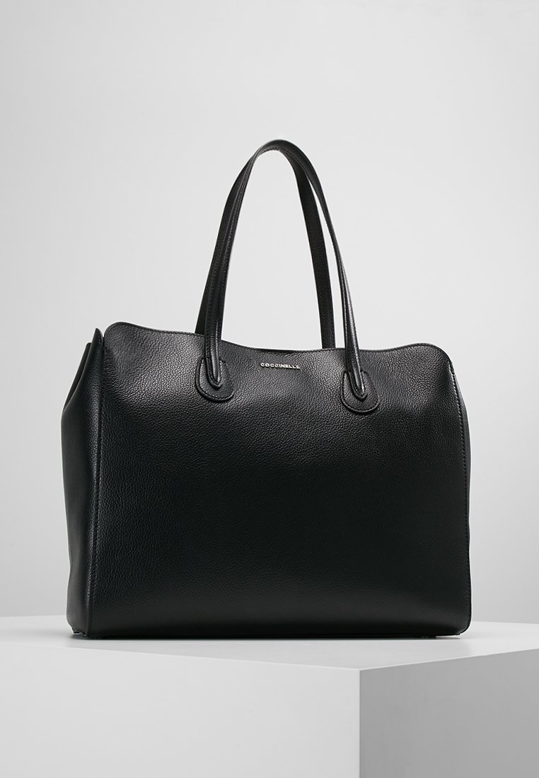 Sac à main en cuir noir avec deux anses, forme structurée et petit logo de marque argenté à l'avant, exposé sur une plateforme blanche.
