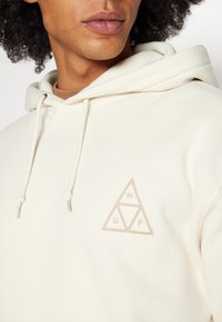 Cremefarvet hoodie med en blød tekstur, der har et trekantet logo i lysebrun på brystet og snore med metalspidser.