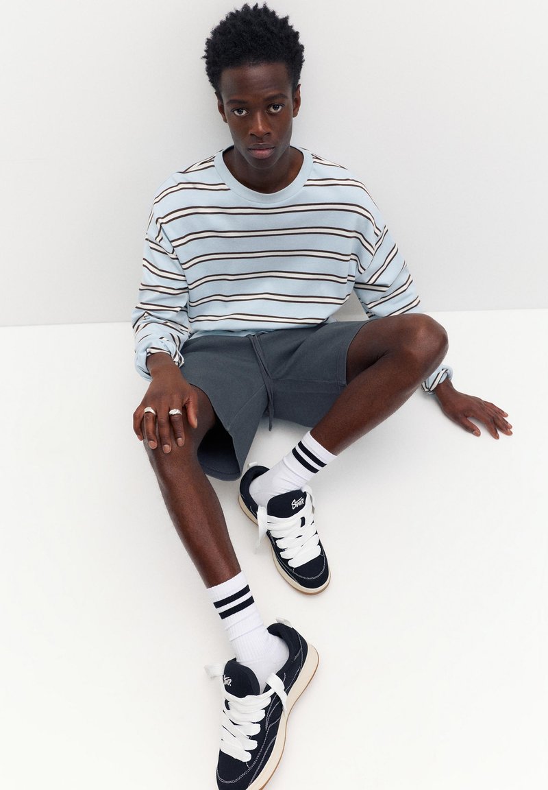 Jeune homme assis par terre portant une chemise rayée bleu clair à manches longues, un short gris, des chaussettes blanches avec des rayures noires et des baskets noires.