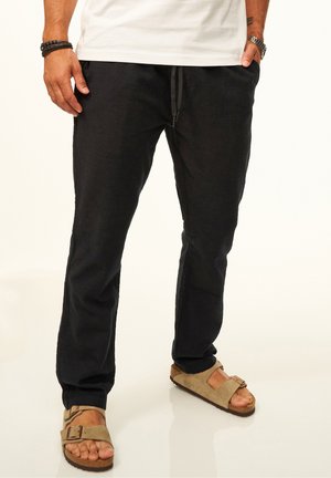 Man wearing black drawstring pants and beige open-toe sandals with a buckle, standing against a white background.