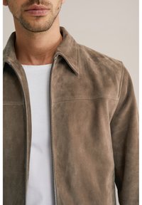 Brūns zamšādas džempera jaka ar apkakli, priekšējo rāvējslēdzēju un gludu tekstūru. Valkāta virs baltas T-krekla, izceļot minimālas dizaina detaļas.