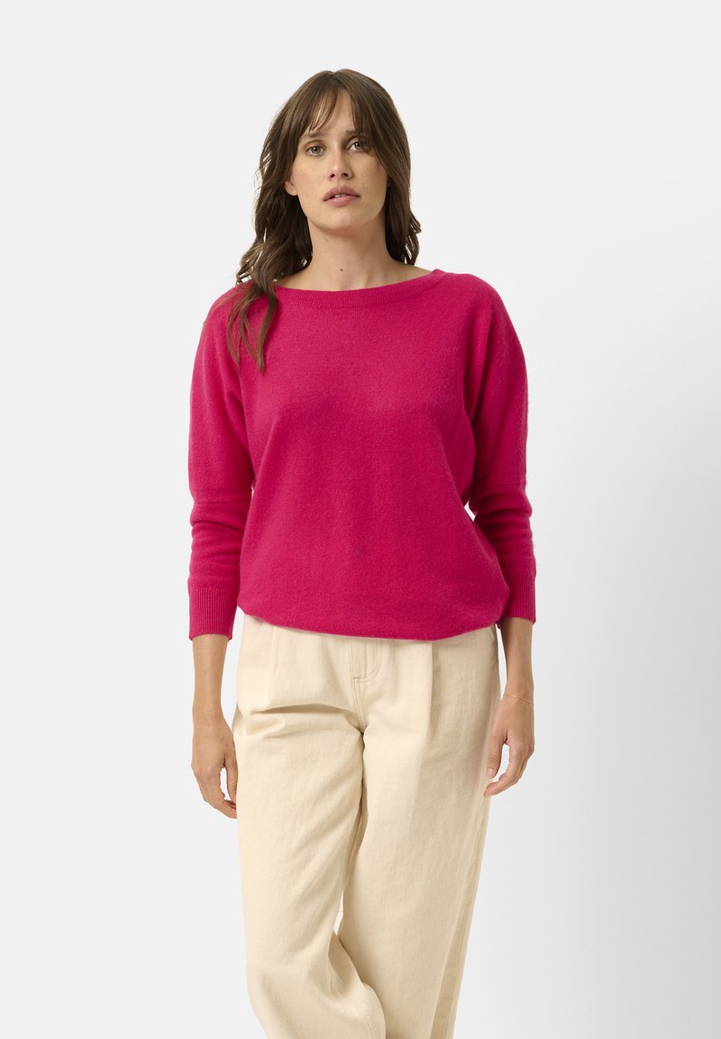 Fuchsia stickad tröja med vid halsringning och långa ärmar, kombinerad med ljusbeiga byxor som skapar en avslappnad silhuett.