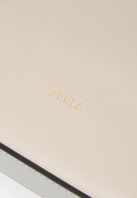 Materiale in pelle beige con il nome del marchio "FURLA" in rilievo color oro. Presenta una texture liscia e una sottile bordatura nera lungo il bordo.