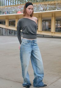 Tânără purtând un top cu o umflare cu dungi, blugi evazați cu aspect decolorat, bentiță neagră și pantofi negri, stând în aer liber lângă o clădire modernă.