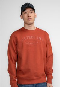 Rode sweatshirt met een gestructureerd "PETROL IND" logo, ronde hals en een patch op de linker mouw. Draagt het met blauwe jeans en een donkere pet.