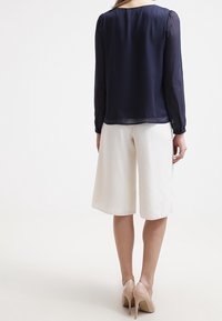 Marineblaue, transparente Bluse mit langen Ärmeln und gerafften Schultern, kombiniert mit weißen, weiten Culottes. Beigefarbene High Heels vervollständigen das Outfit.