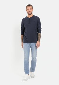 Marineblaues Langarm-Henley-Shirt mit Knopfleiste, kombiniert mit hellblauen Jeans und weißen Sneakern. Stylisch mit sichtbaren Tattoos an den Armen.