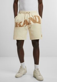 Cream-colored shorts with a textured brown 'KANDY' logo, featuring an elastic waistband and side pockets. Paired with cream sneakers.