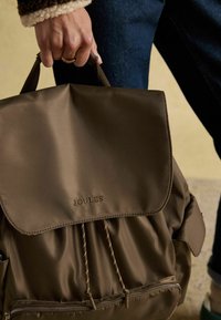 Rjava najlonska nahrbtnik z zgornjim pokrovom, zapiranjem na vrvico in sprednjim žepom z zadrgo. Ima vtisnjen logotip "JOULES" in teksturirano tkanino.