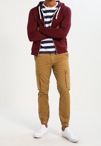 Sudadera con cremallera color burdeos sobre una camiseta de rayas, combinada con pantalones cargo color caqui y zapatillas blancas. Los pantalones tienen bolsillos laterales y puños elásticos.