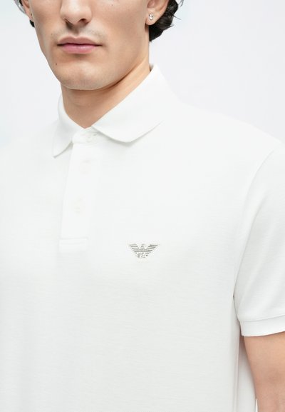 Biele polokošeľa s jemnou textúrou, má klasický golier, tri gombíky a malý strieborný vyšívaný logotyp na prsiach.