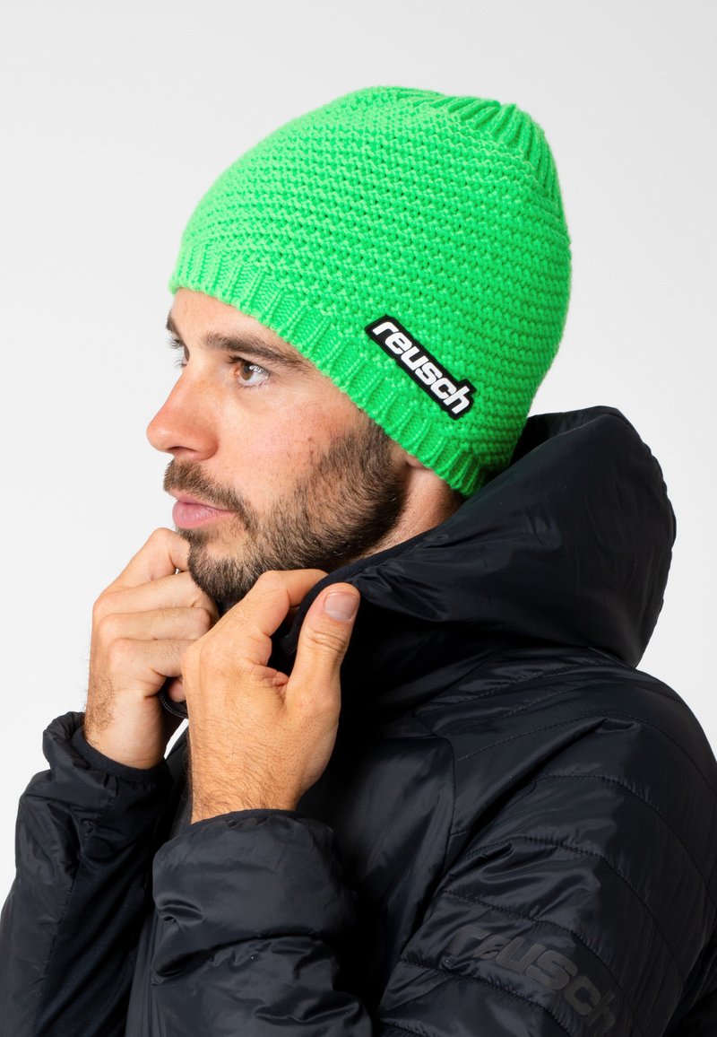Green knitted beanie with textured pattern and a black Reusch logo patch, worn by a person in a black jacket, facing to the side.