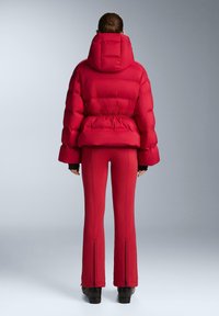 Piros tollkabát magas gallérral, rugalmas derékrésszel és nagy kapucnival, hozzá illeszkedő piros nadrággal, amelyen cipzárok vannak a bokáknál.