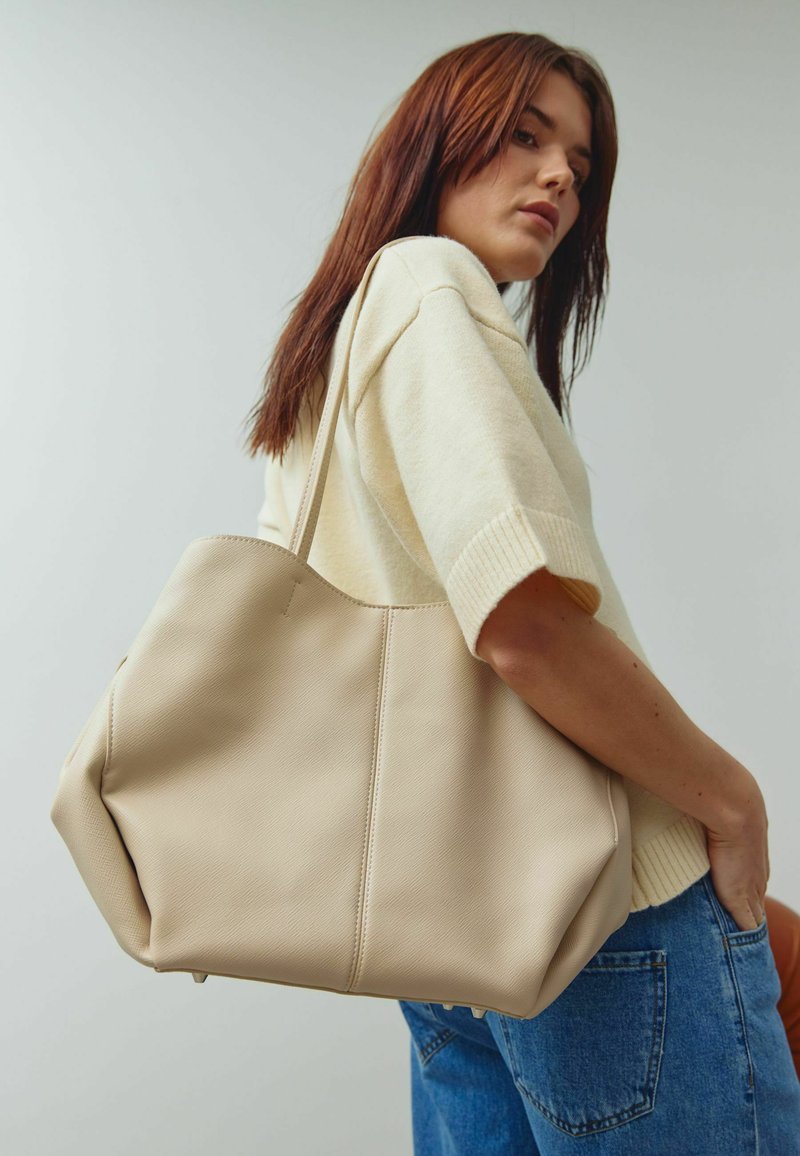 Geantă tote din piele bej deschis, cu o textură moale, având cusături laterale și un design spațios. Purtați pe umăr, ținută cu o singură mână.