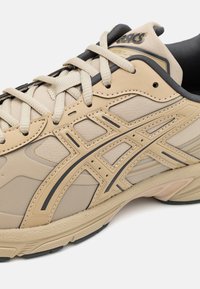 Chaussure de course beige Asics avec logo noir, lacets beiges et semelle intermédiaire texturée, montrée en gros plan de côté.