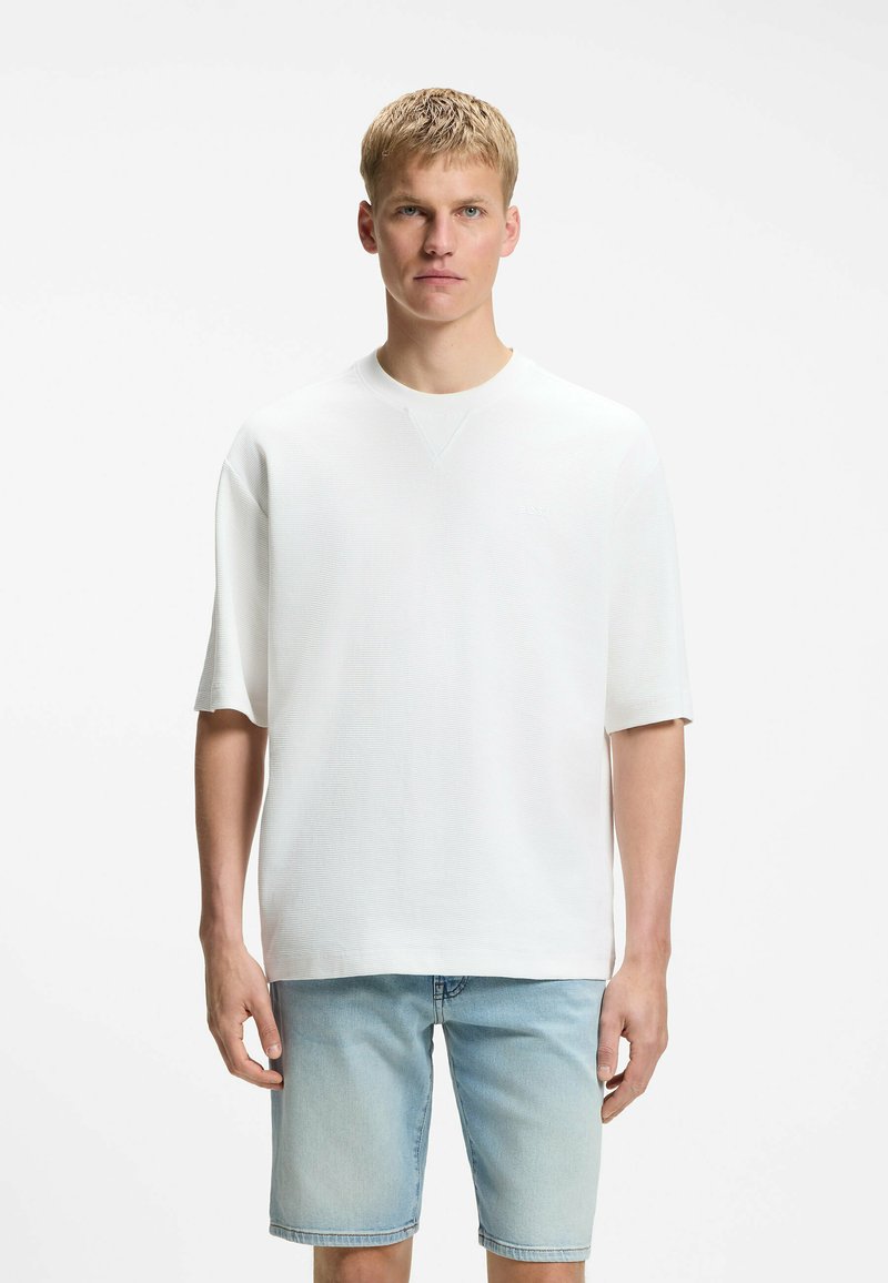 Homme blond portant une chemise blanche à manches courtes ample et un short en jean bleu clair, debout devant un fond blanc uni.