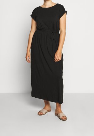 Frau trägt ein schwarzes Midi-Kleid mit kurzen Ärmeln und Taillengürtel sowie beigen Slip-On-Sandalen, steht vor weißem Hintergrund.