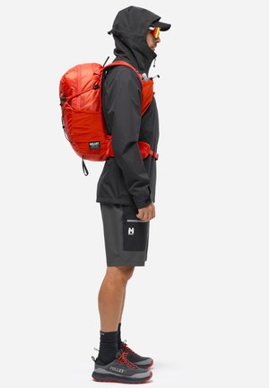 Homme en tenue de plein air avec veste à capuche noire, short gris, sac à dos rouge, casquette, lunettes de soleil et chaussures de randonnée, debout de profil sur fond blanc.