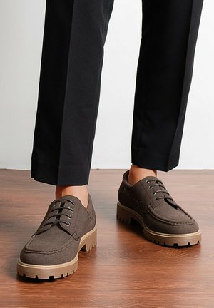 Chaussures décontractées marron à lacets avec semelles épaisses beiges portées avec un pantalon noir sur un sol en bois.
