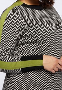 Black and white patterned sweater featuring a herringbone design, with green accents on the shoulders and cuffs. Soft texture, relaxed fit.