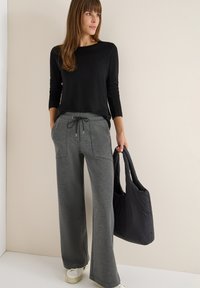 Schwarzes Langarmoberteil, graue weit geschnittene Hose mit Taschen und Kordelzug, schwarze gesteppte Tragetasche, weiße Sneakers. Schlichtes, modernes Design.