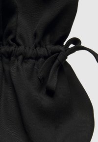 Pochette en tissu noir avec surface texturée, haut froncé et fermeture à cordon. Comporte un matériau doux et des liens lisses et ronds.