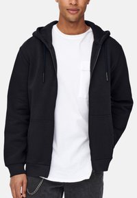 Schwarze Zip-Hoodie mit Kapuze zum Zusammenziehen, vorderen Taschen und gerippten Bündchen, getragen über einem weißen Shirt. Der Stoff wirkt weich und texturiert.