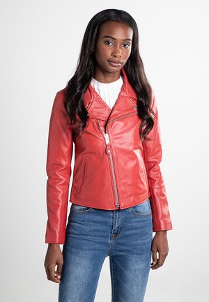 Jeune femme aux longs cheveux portant une veste en cuir rouge zippée sur un haut blanc et un jean bleu, debout devant un fond clair.