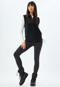 Mustaka fleece-vesti, joka on varustettu vetoketjulla, yhdistettynä mustiin leggingseihin ja polvipitkiin mustiin saappaisiin. Malli seisoo neutraalissa ympäristössä.