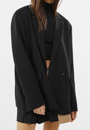 Frau trägt einen übergroßen schwarzen Blazer, ein schwarzes Crop-Top und einen zweifarbigen schwarz-beigen gestreiften Rock und steht vor einem schlichten Hintergrund.