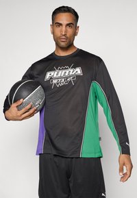 Μακρυμάνικη μαύρη αθλητική μπλούζα με πράσινες, μωβ και μαύρες πλευρικές λεπτομέρειες, που φέρει το λευκό λογότυπο "PUMA" και κρατάει μια υφασμάτινη μπάλα μπάσκετ.