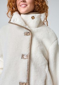 Vit fleecejacka med hög krage, kontrasterande beige kant, och två runda knappar. Mjuk textur med en mysig känsla.