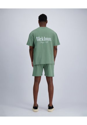 Mann steht mit dem Rücken zum Betrachter, trägt grüne Shorts und passendes T-Shirt mit dem Schriftzug „Blckbnns Design Dept“, schwarz-weiße Sneakers.