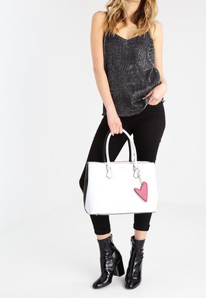 Kvinde iført sort ærmeløs top og bukser, der holder en hvid håndtaske med lyserød hjertevedhæng, iført sorte skinnende ankelstøvler.